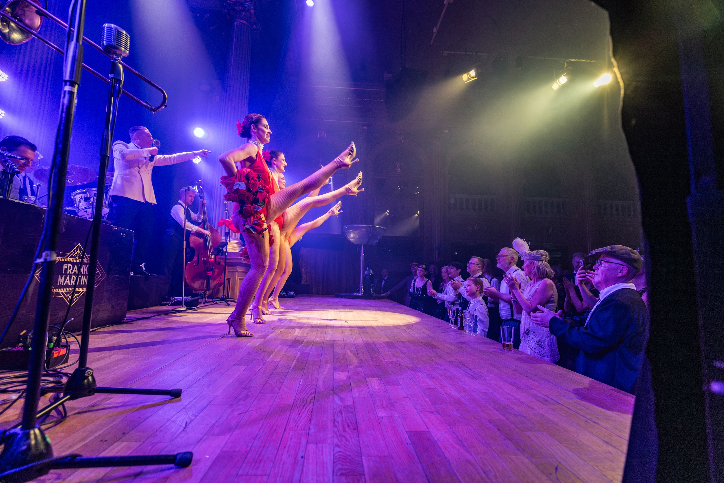 Musicians perform on stage under dramatic purple lighting while an audience watches from the side at an intimate concert venue.