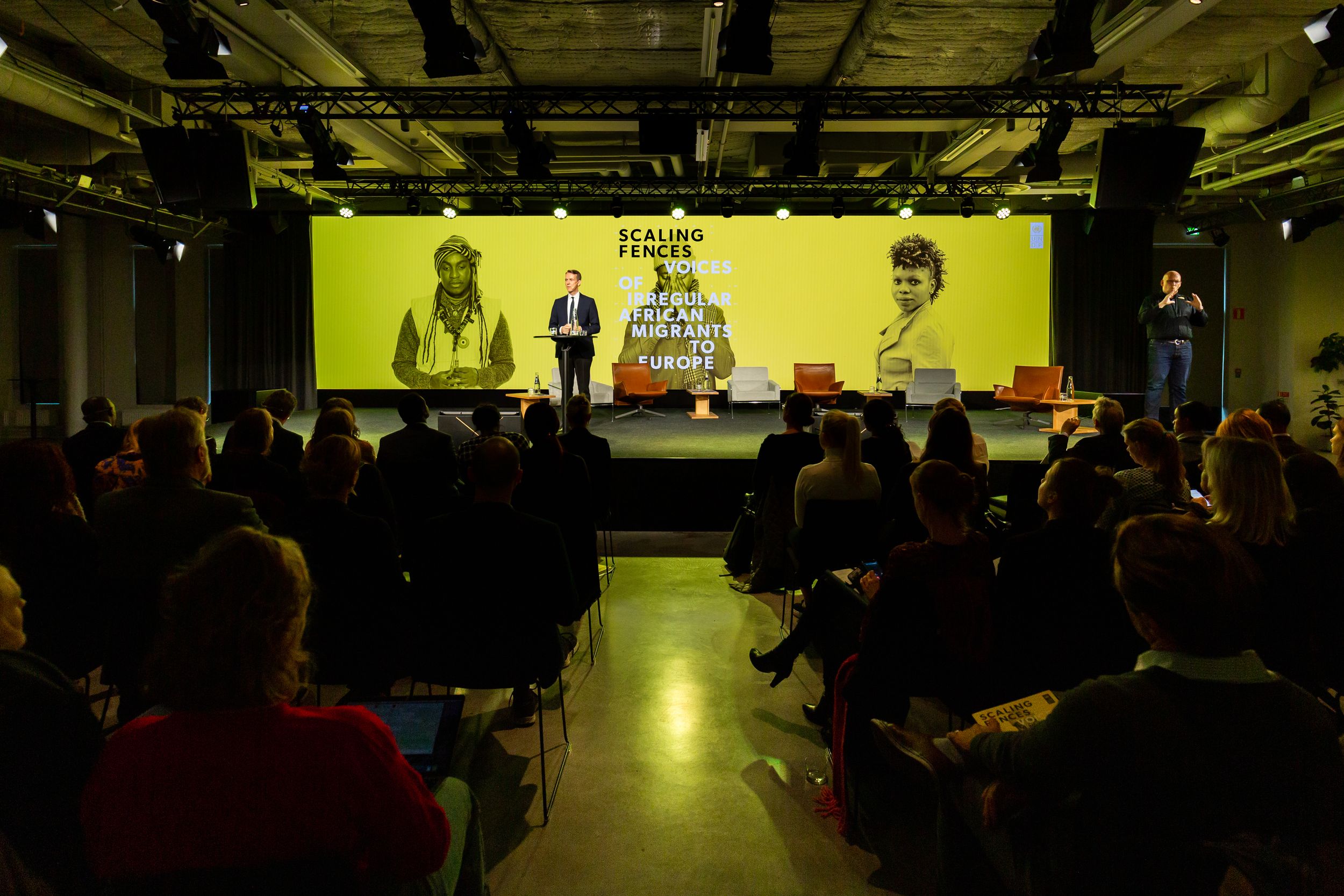 Eventfotography of an audience watching a presentation on a large screen in dimly lit conference space with industrial ceiling.