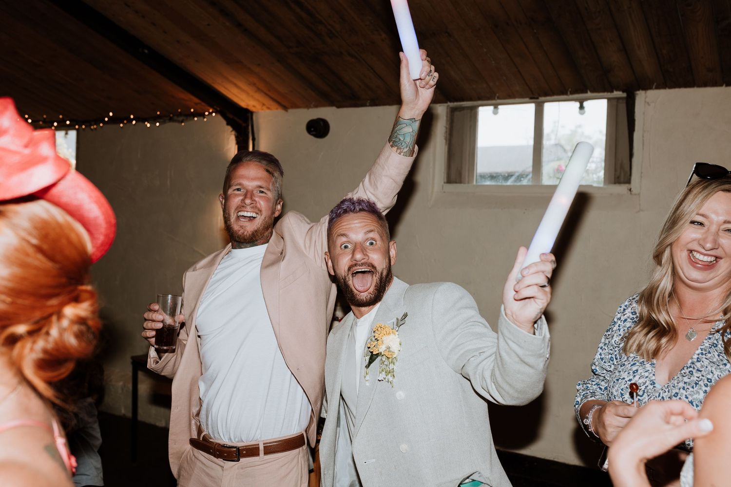 Two joyful men celebrating at a wedding reception, raising their arms with paper rolls in a rustic venue.