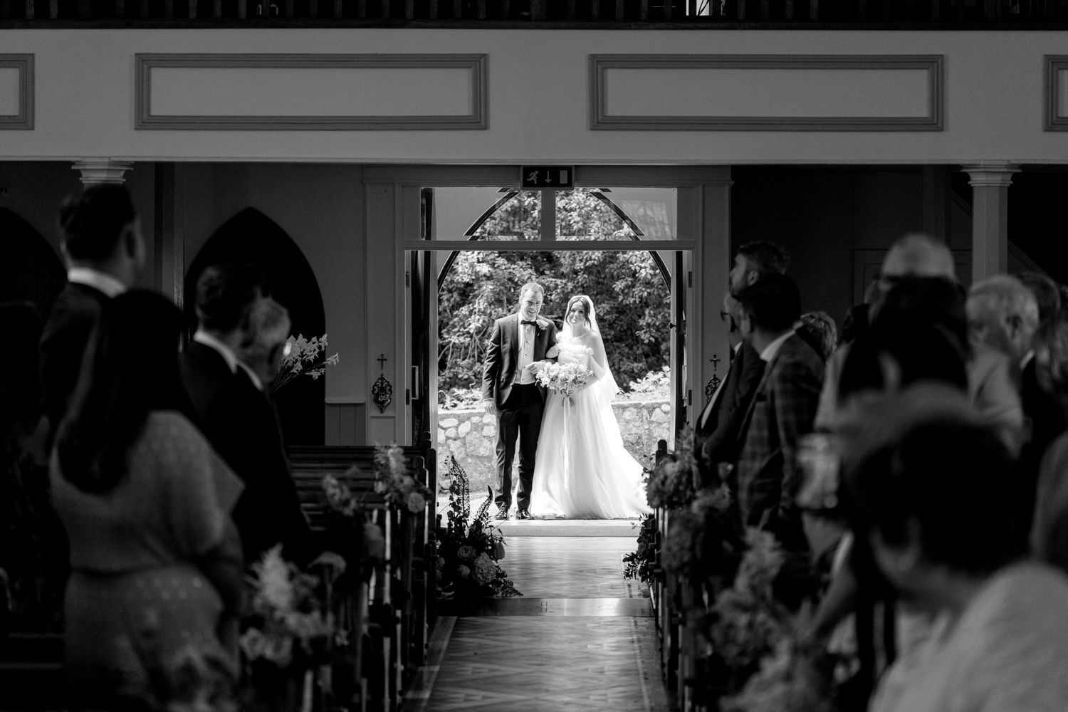 Black and white photo of a bride walking down a hallway in castle-like setting with onlookers visible in background.
