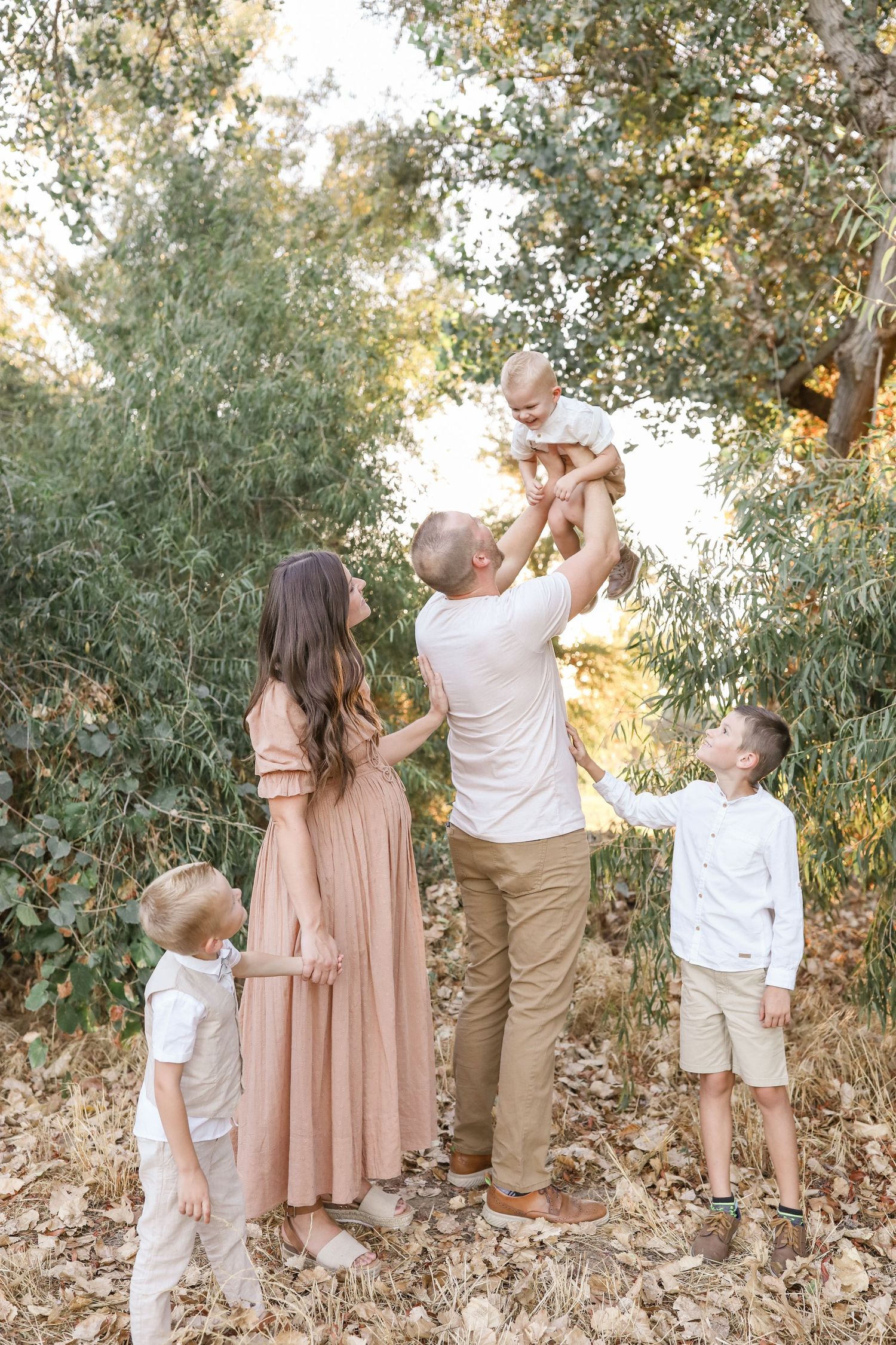 Family of five enjoying outdoor portrait session, father lifting baby while children gather around mom in blush dress.