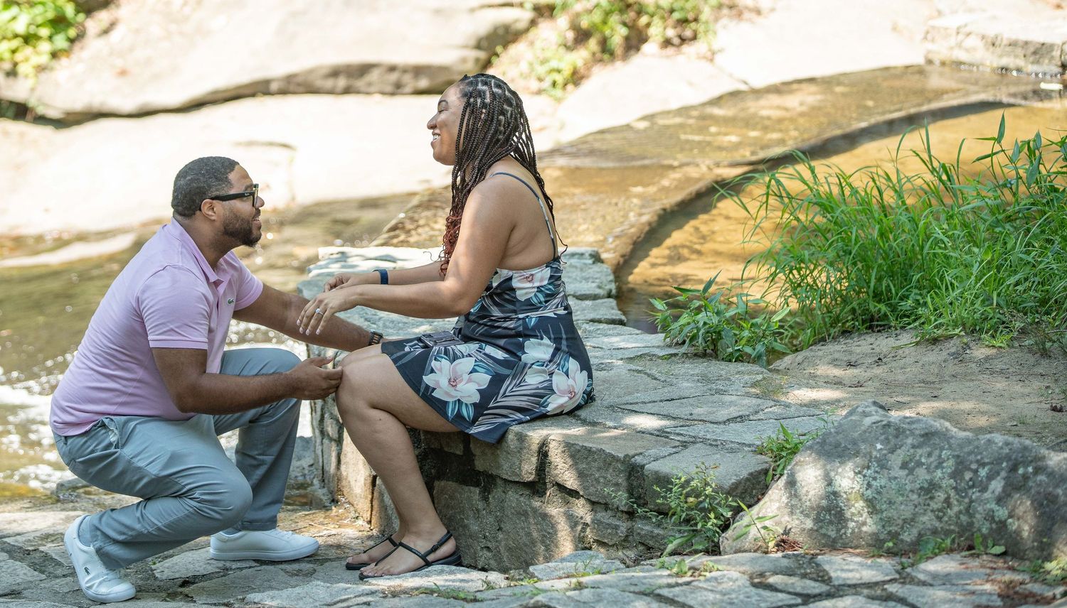 Man kneeling before a woman seated on rocks near a stream in a lush park, appearing to propose marriage.