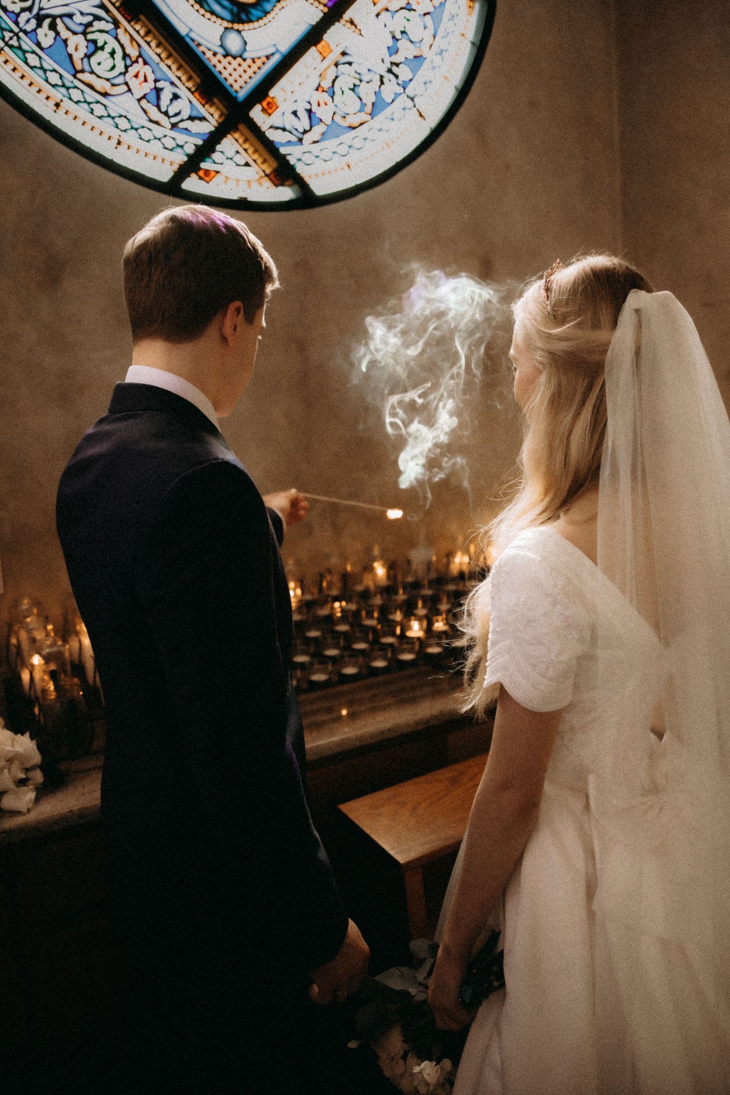 Bride and groom lighting candles in a church with stained glass window, smoke rising from a match.