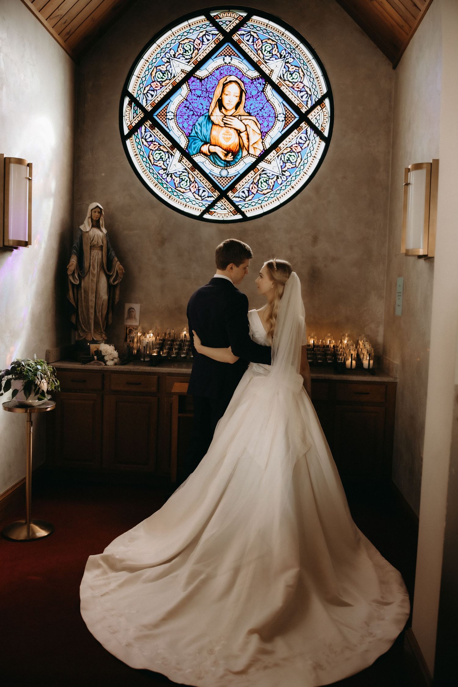Bride and groom share romantic moment in chapel with stunning stained glass Madonna window and candlelit altar.