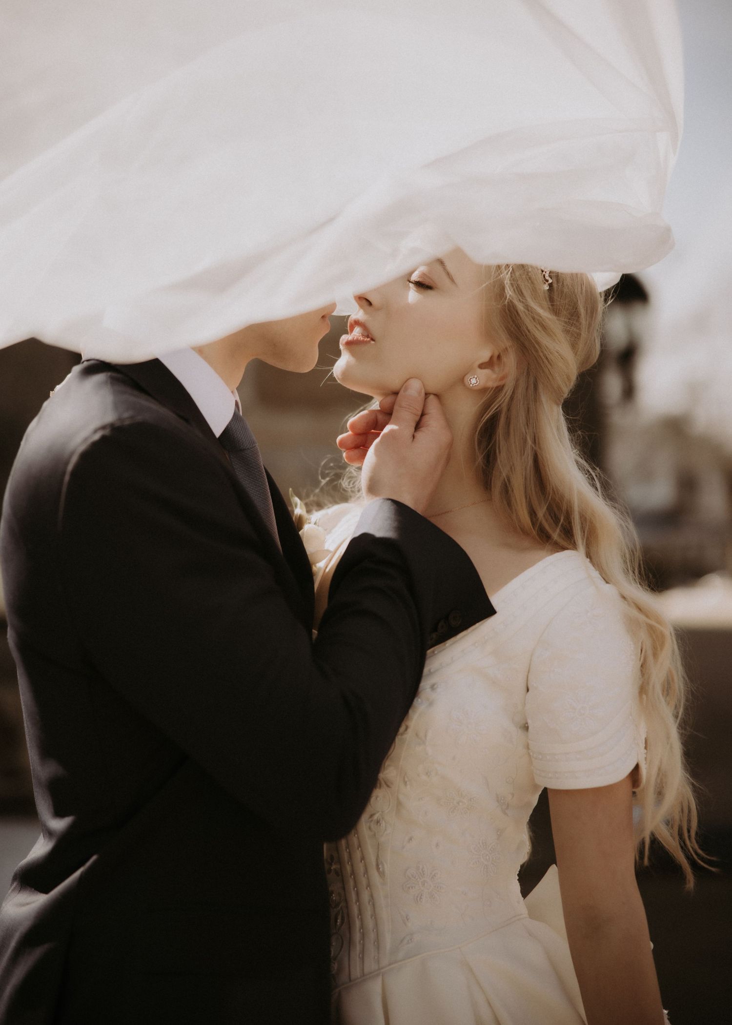 A groom tenderly holds his brides chin under a flowing veil in a romantic outdoor wedding portrait.