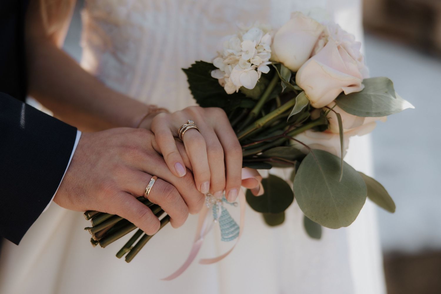 Newlyweds hold hands together over a bridal bouquet of white roses, showing off their wedding rings.