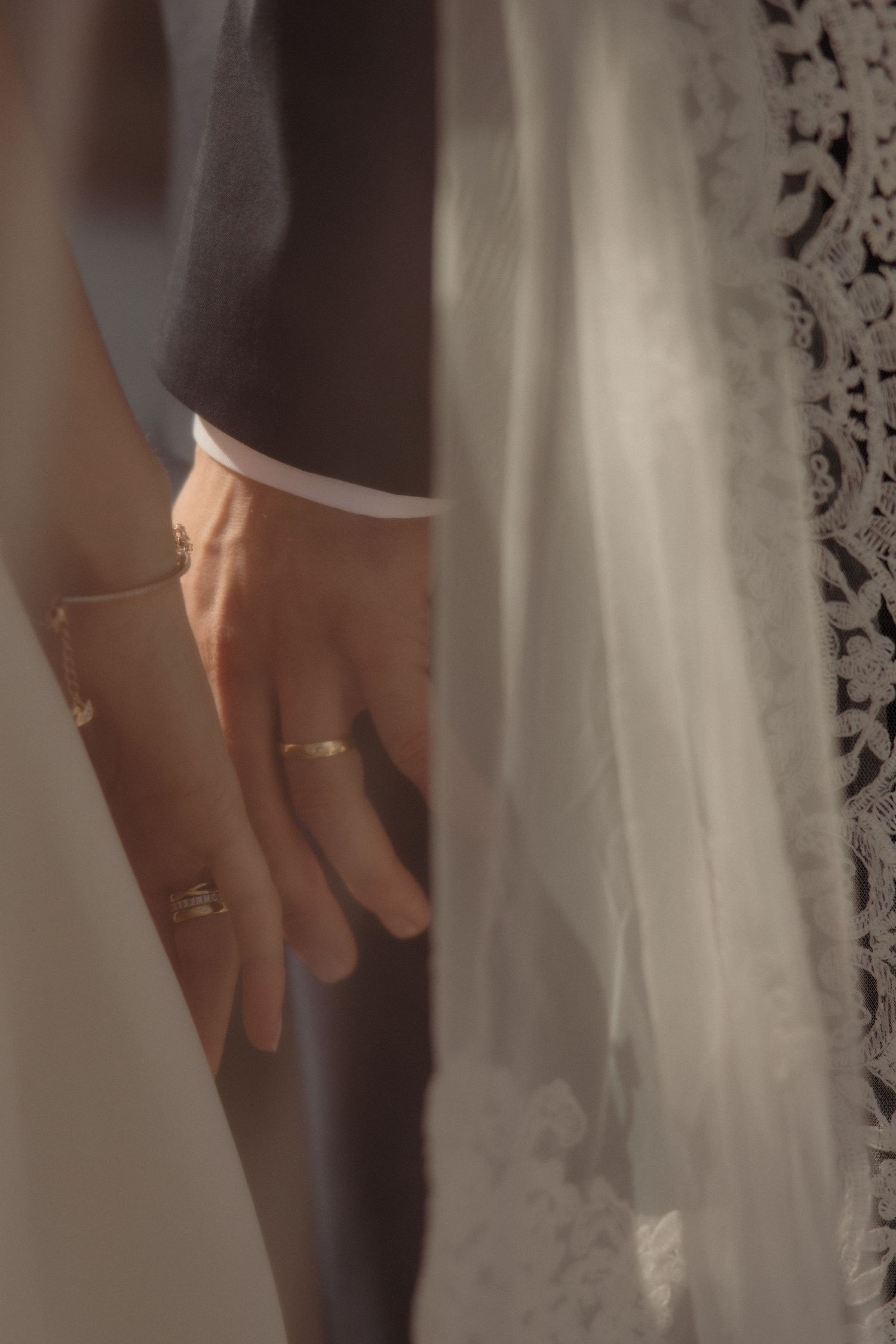 Close-up of bride and groom holding hands, showcasing wedding rings and bracelet with soft light and lace veil.