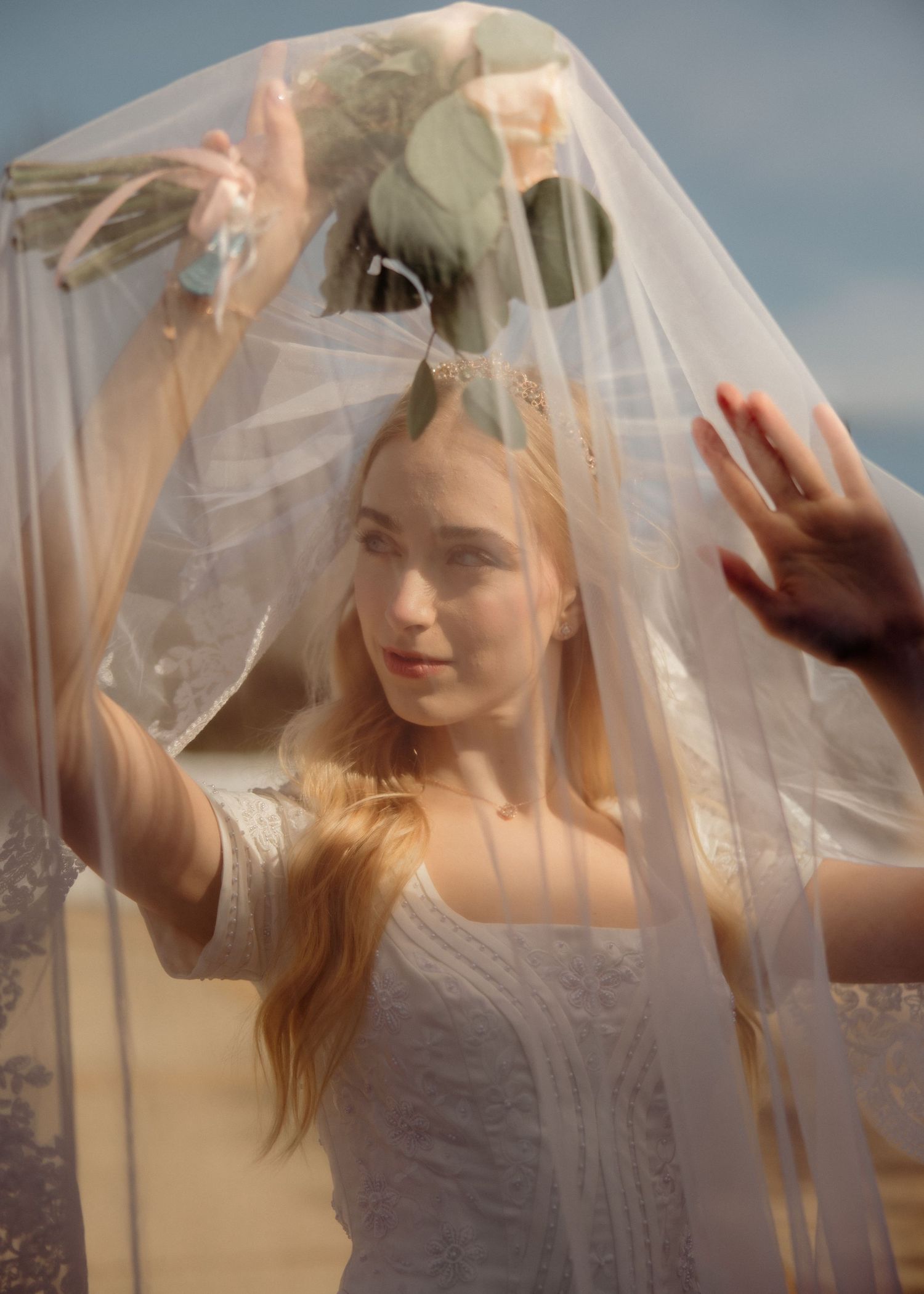Bride in lace wedding dress holding bouquet under flowing veil in soft natural light.