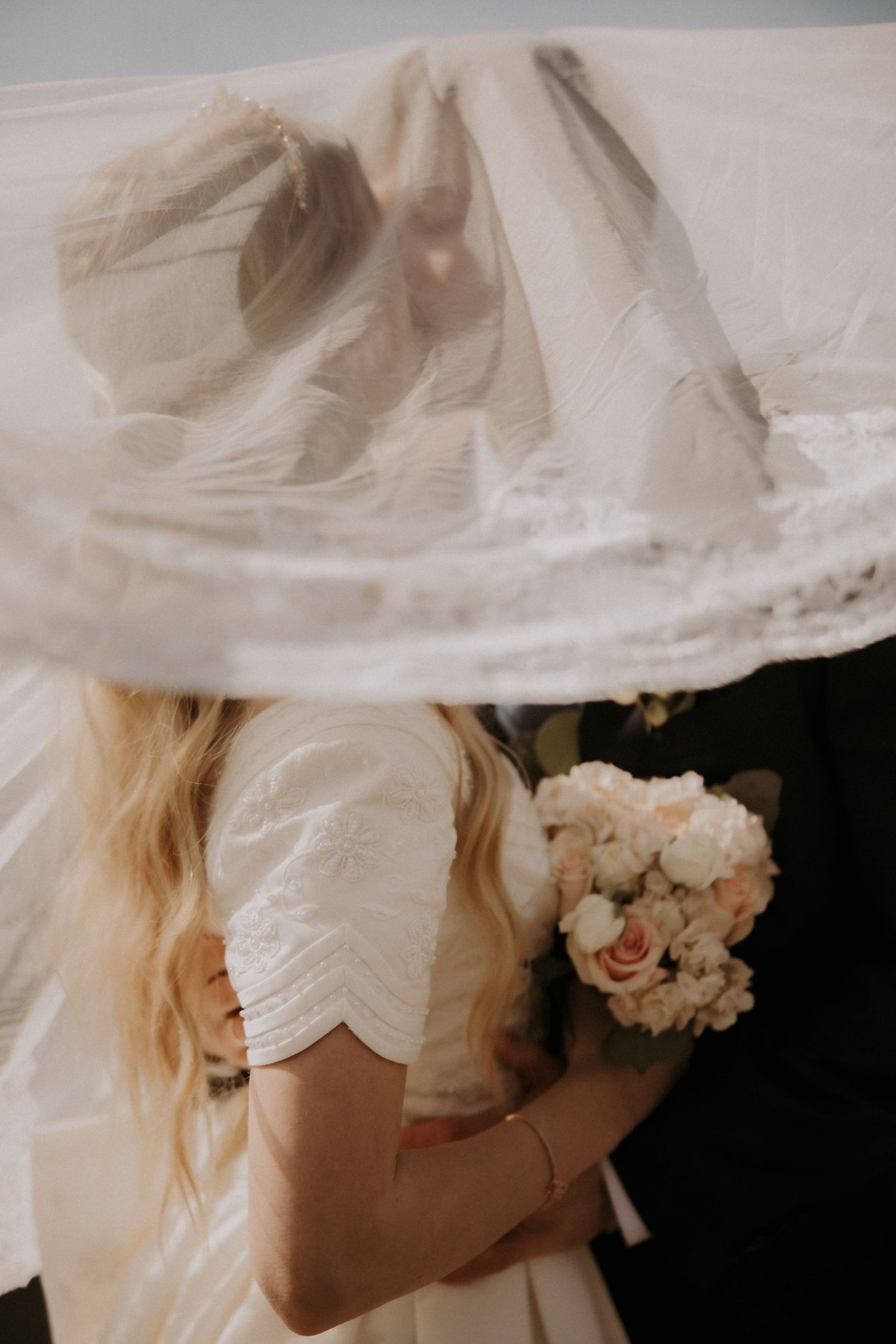 Bride holding white and pink bouquet embracing groom under billowing wedding veil.