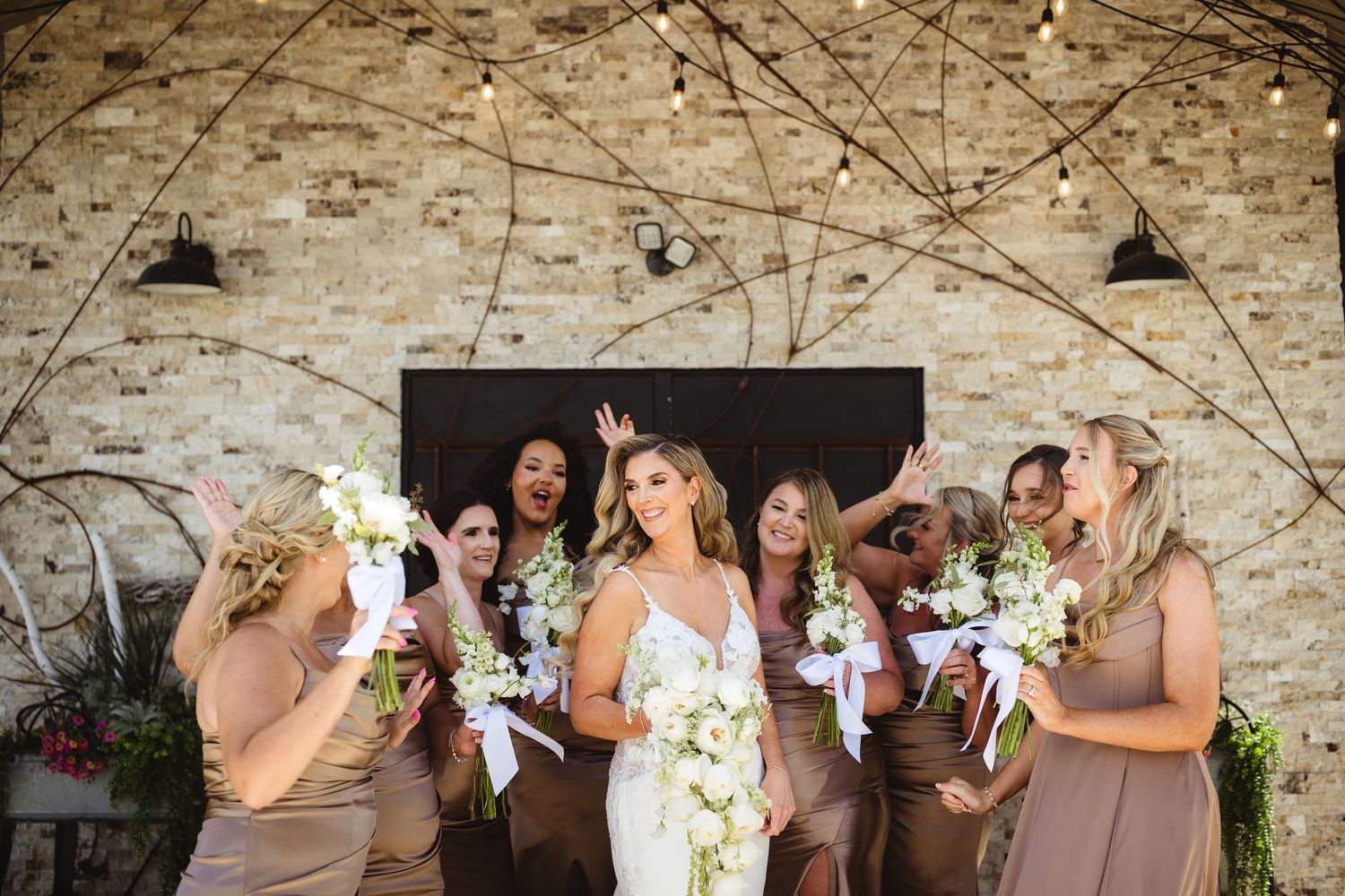 Bride in white lace gown celebrates with bridesmaids in taupe dresses holding white bouquets at rustic venue.