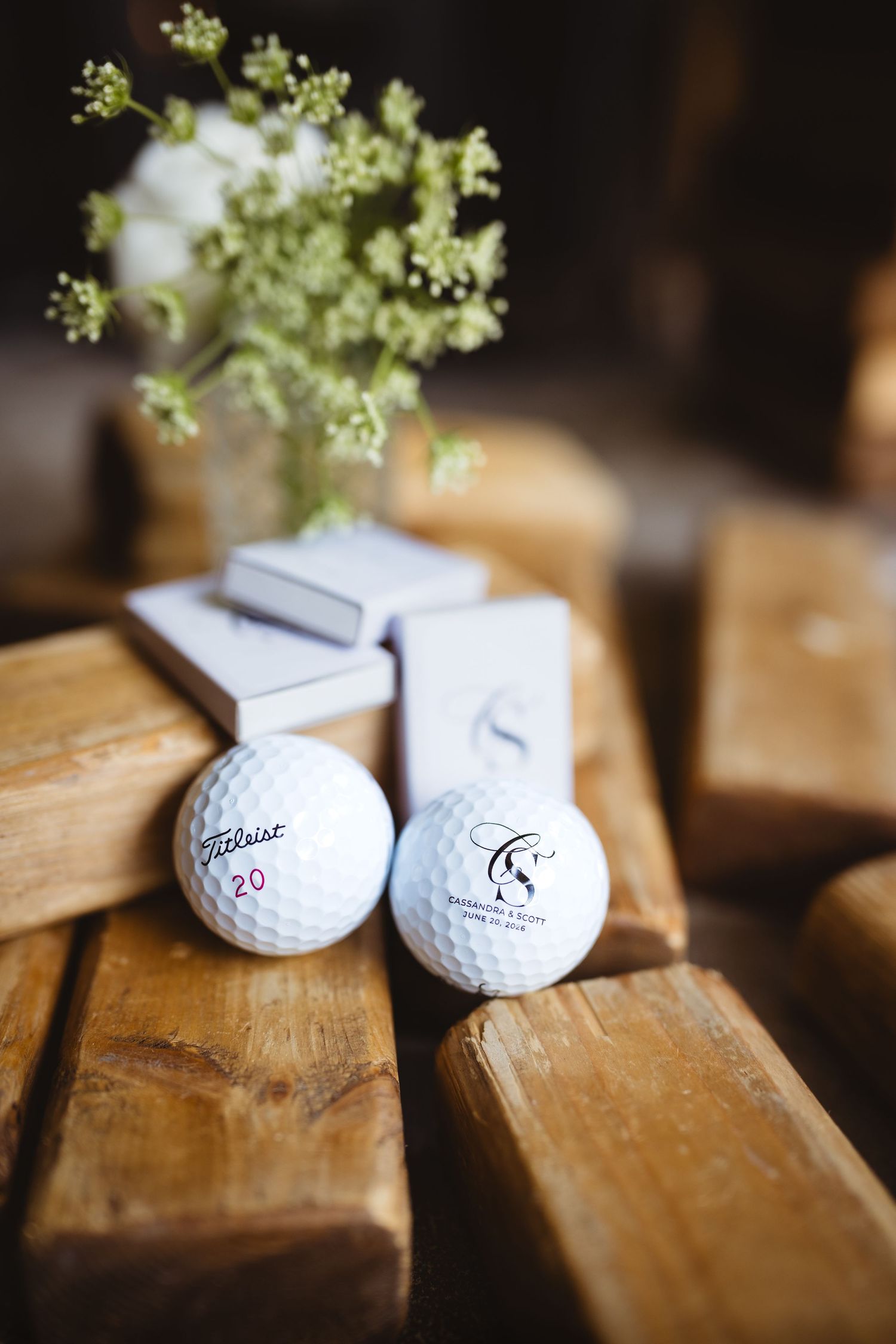 Two Titleist golf balls on rustic wooden surface, one personalized with monogram, beside white flowers and matchbox.