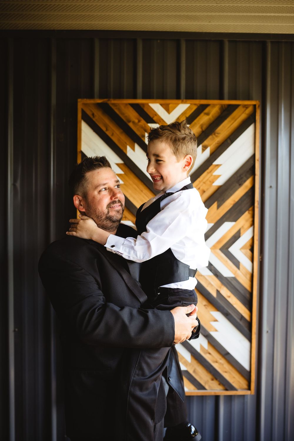 A man in a black suit holds a laughing boy in a white shirt and vest, posing before a geometric wood art piece.
