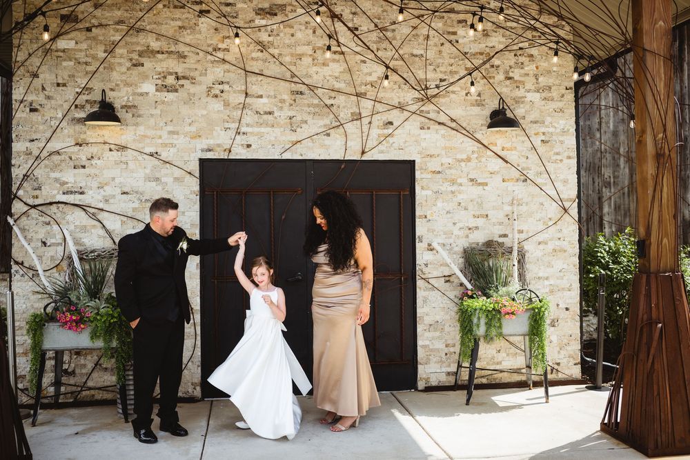 A couple twirls a flower girl in a white dress at a rustic wedding venue with string lights and brick walls.