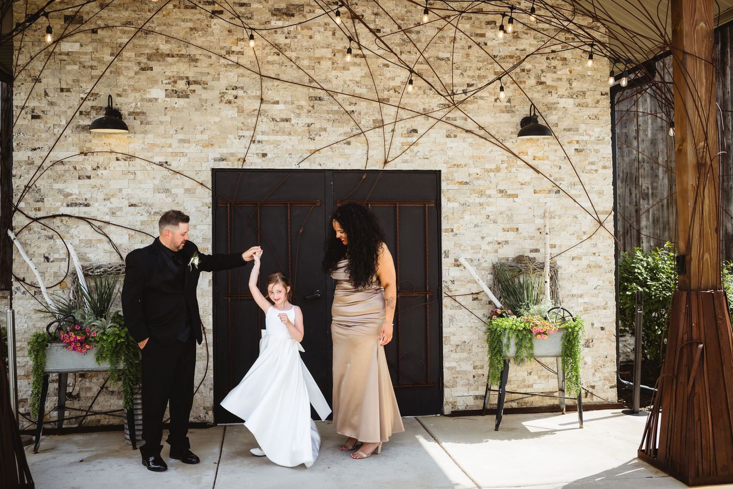 A couple twirls a flower girl in a white dress at a rustic wedding venue with string lights and brick walls.