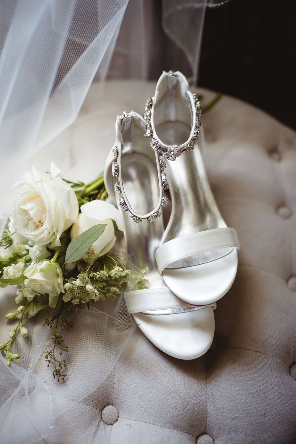 Elegant white bridal heels with crystal ankle straps beside a white rose bouquet on a tufted chair.