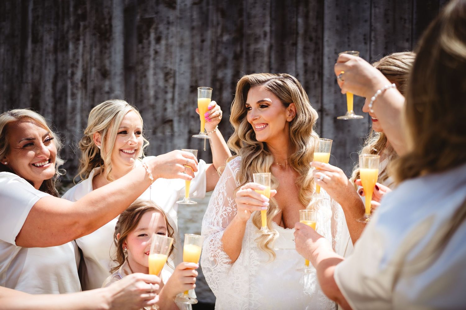 Bride in lace gown toasting champagne with bridesmaids in white robes at rustic barn wedding celebration.
