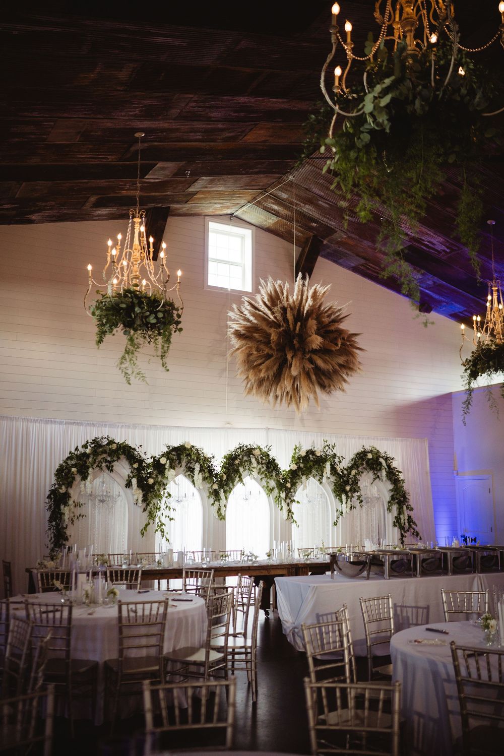 Elegant barn wedding reception with chandelier greenery, floral arch backdrop, white draping, chiavari chairs, and soft purple uplighting.