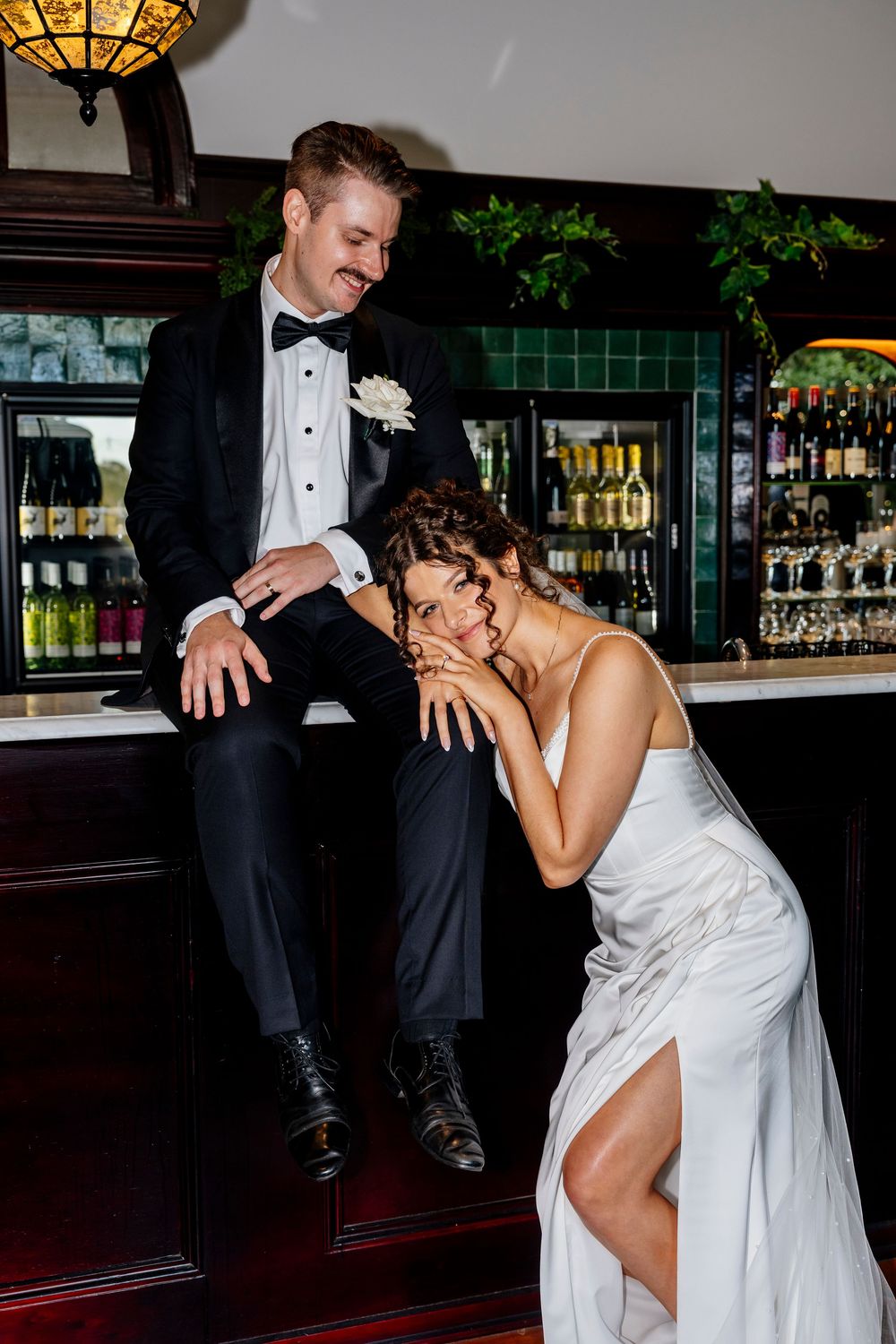 Bride in white gown leans on bar counter beside groom in black tuxedo at elegant wedding venue in queensland
