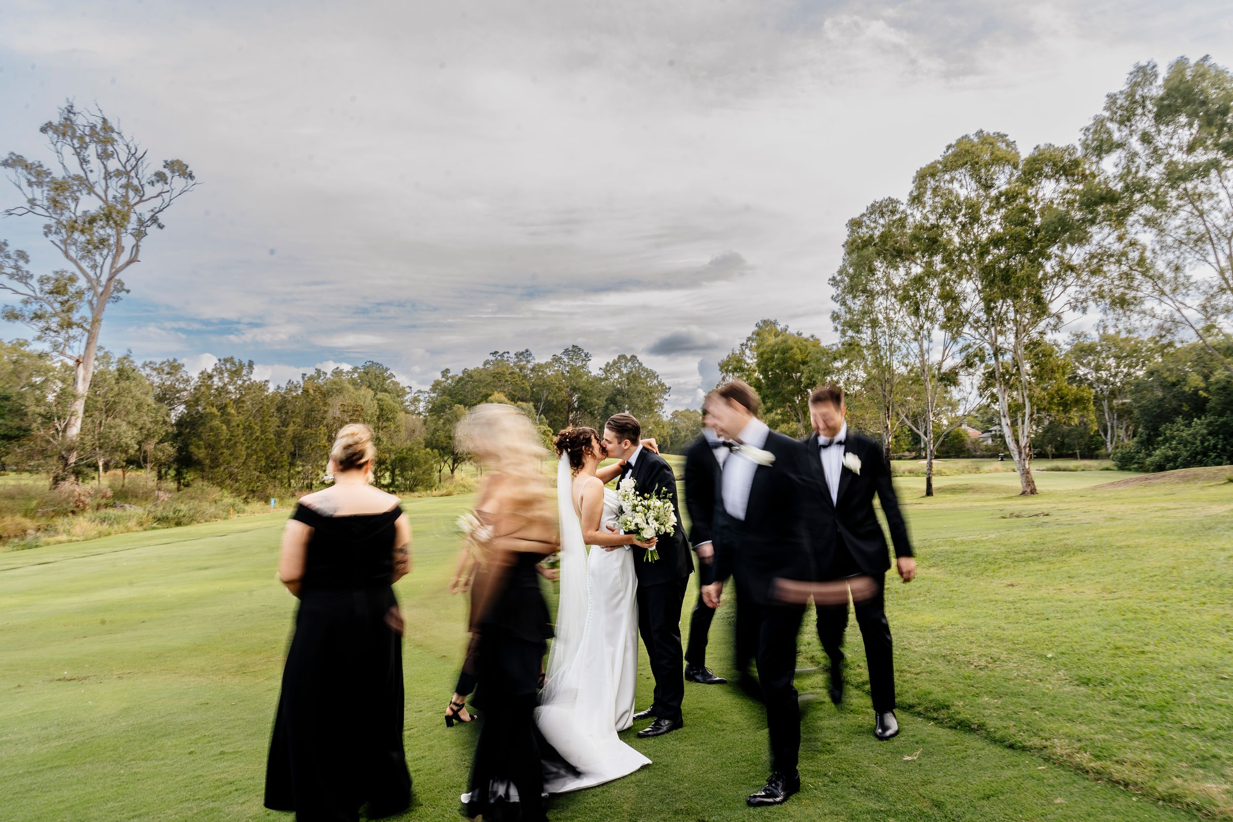 Wedding party in black attire and bride in white gown celebrating on lush green golf course with trees under cloudy sky.
