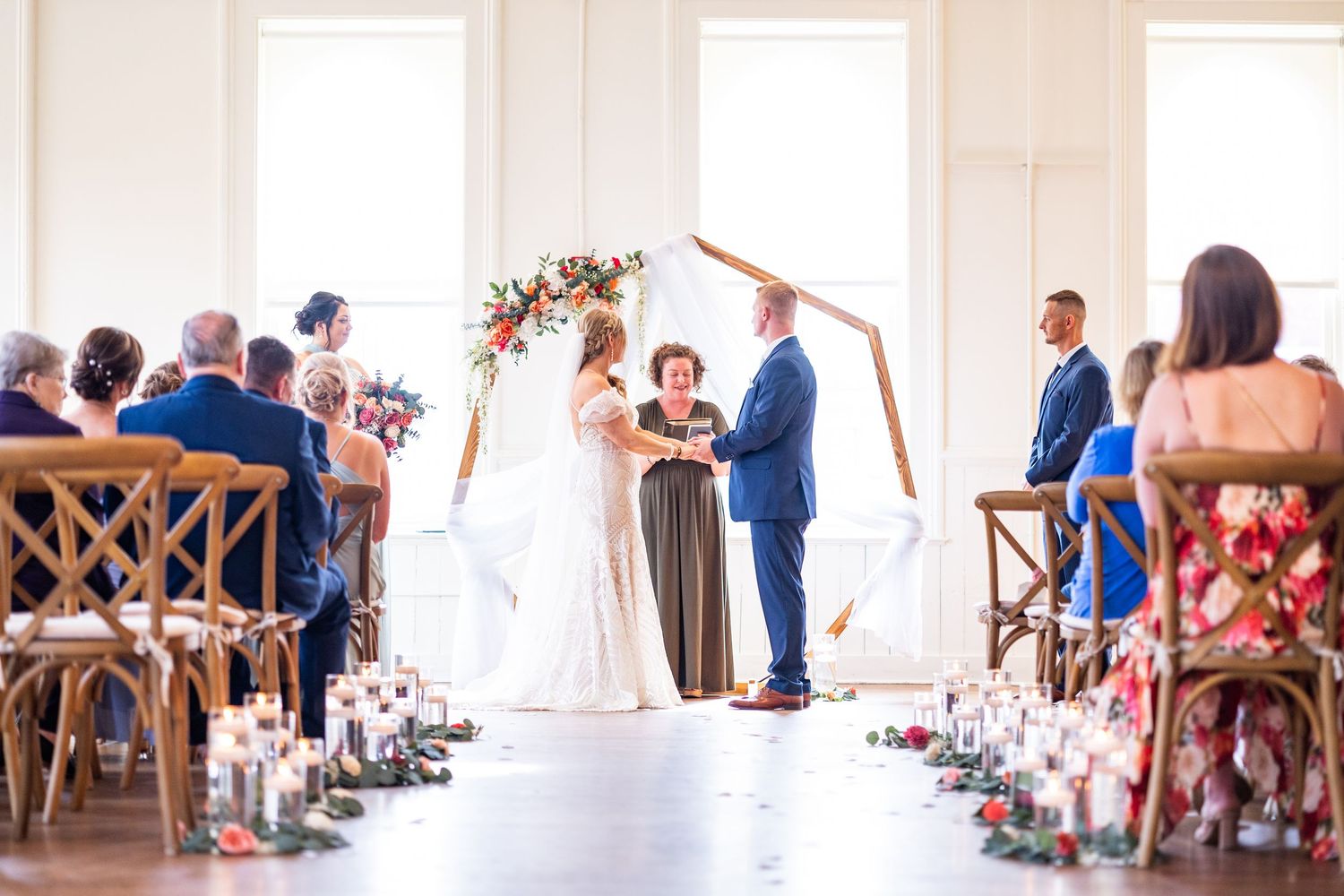 Elegant indoor wedding ceremony at the Alcove Cincinnati with couple at floral arch, candles lining aisle, and guests in wooden chairs.
