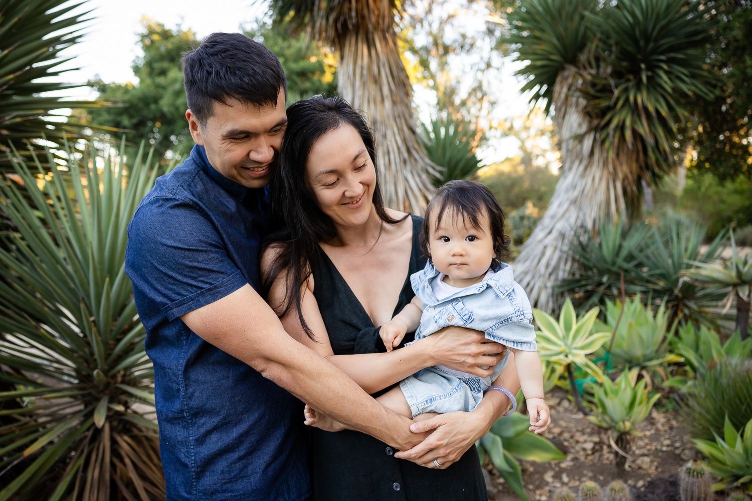 A happy family of three poses outdoors, parents smiling at their baby boy amid garden greenery in the Palo Alto cactus garden at Stanford.