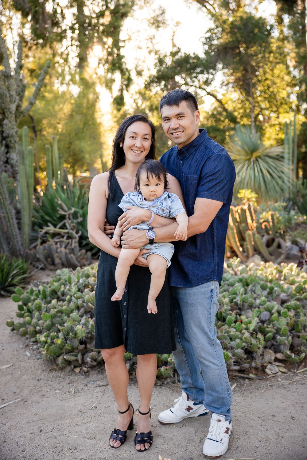 Happy family of three posing in a lush garden, parents holding baby boy among cacti and succulents.