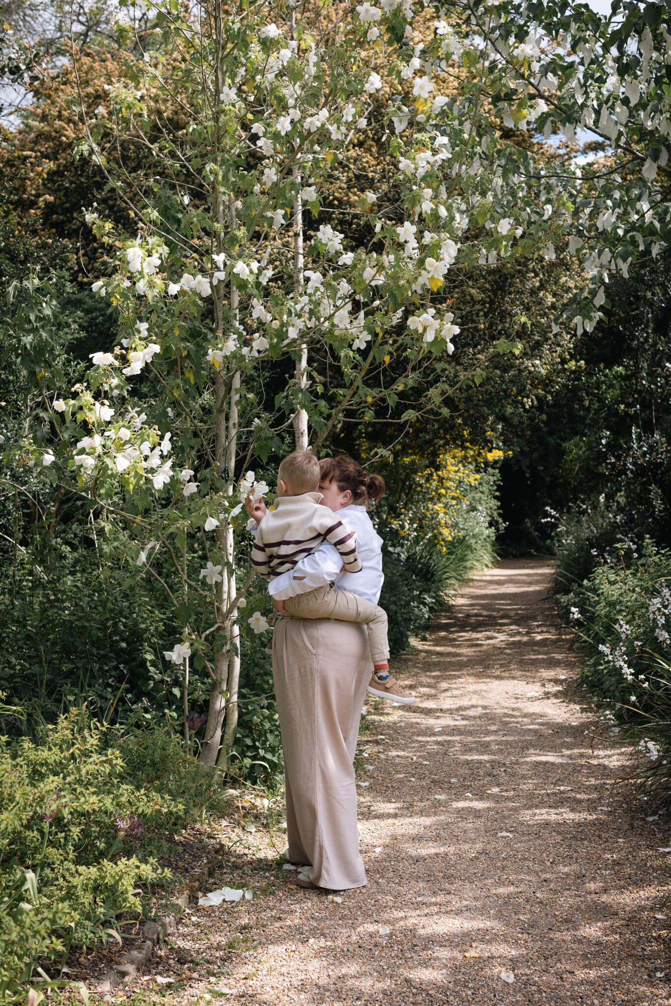 A mother holding her baby while standing on a garden path surrounded by lush greenery and white flowering trees, photographed in Sussex