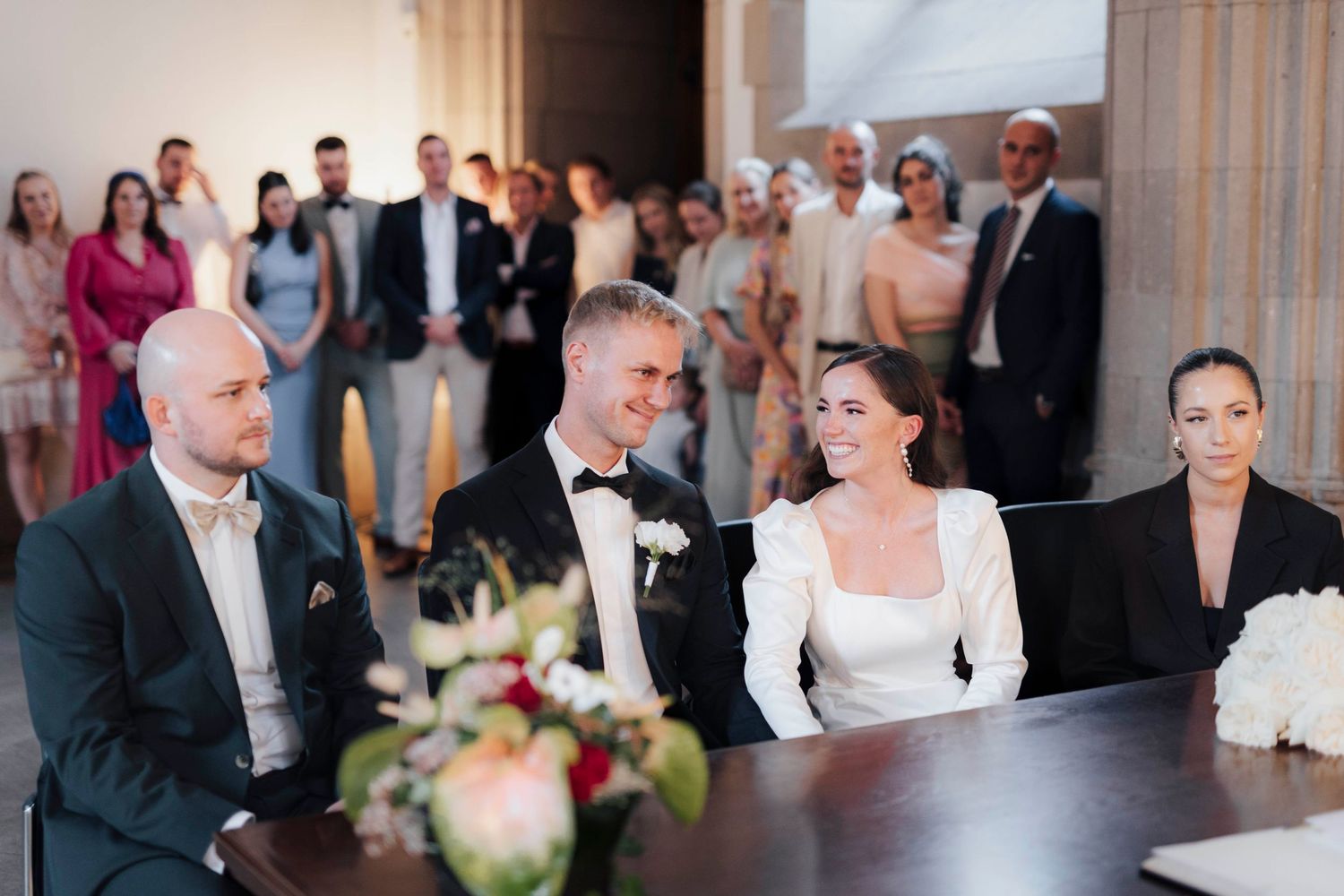 Bride and groom smile at wedding ceremony table, guests standing behind them in elegant venue.