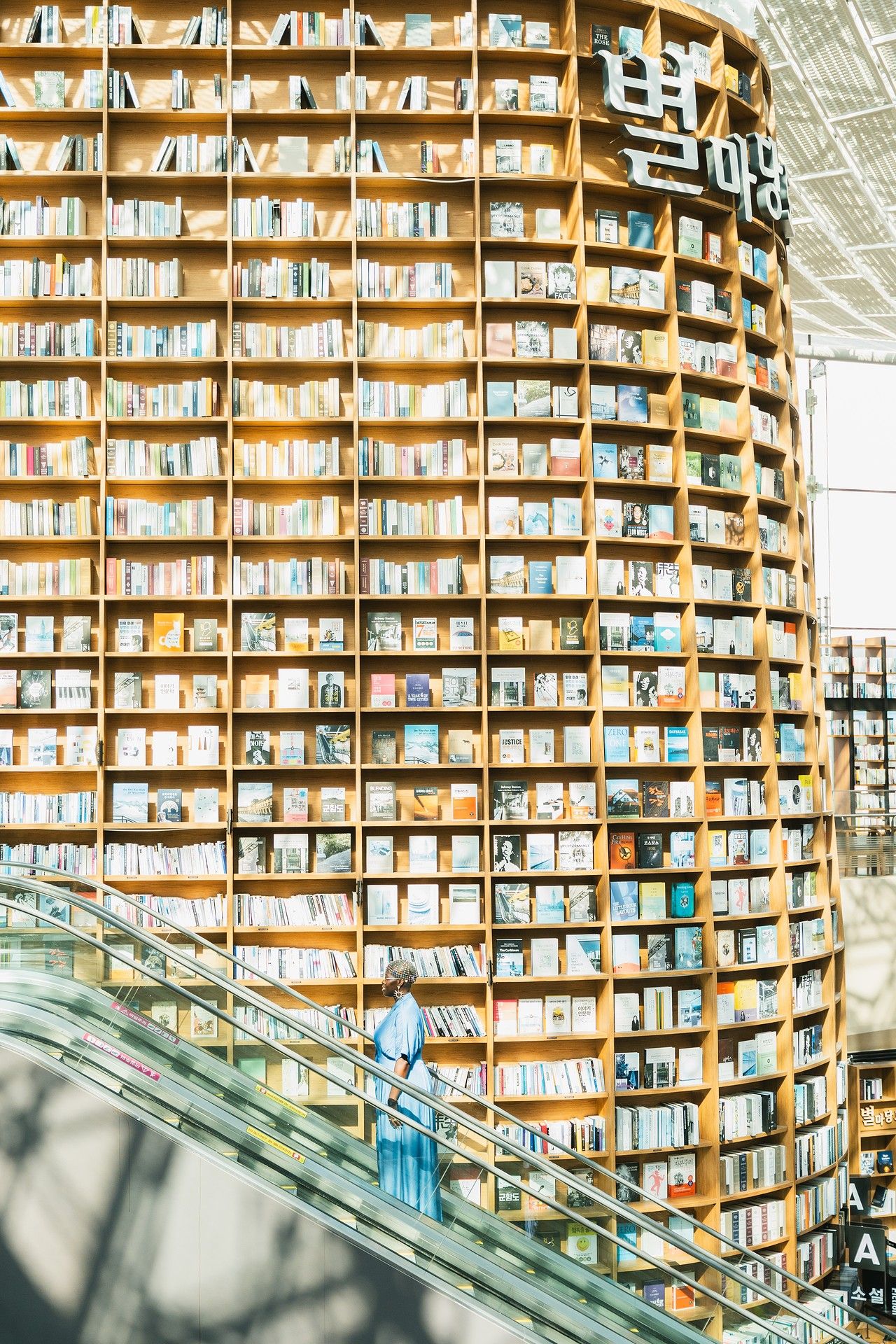Woman on escalator beside a massive curved bookshelf filled with books in a modern Korean library.