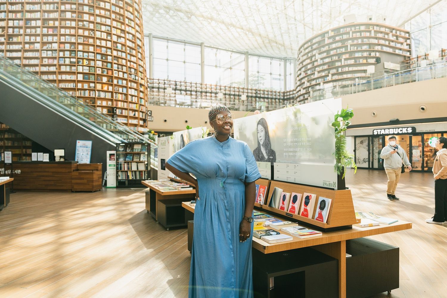Woman in blue dress standing in a modern library atrium with curved bookshelves and a Starbucks in the background.