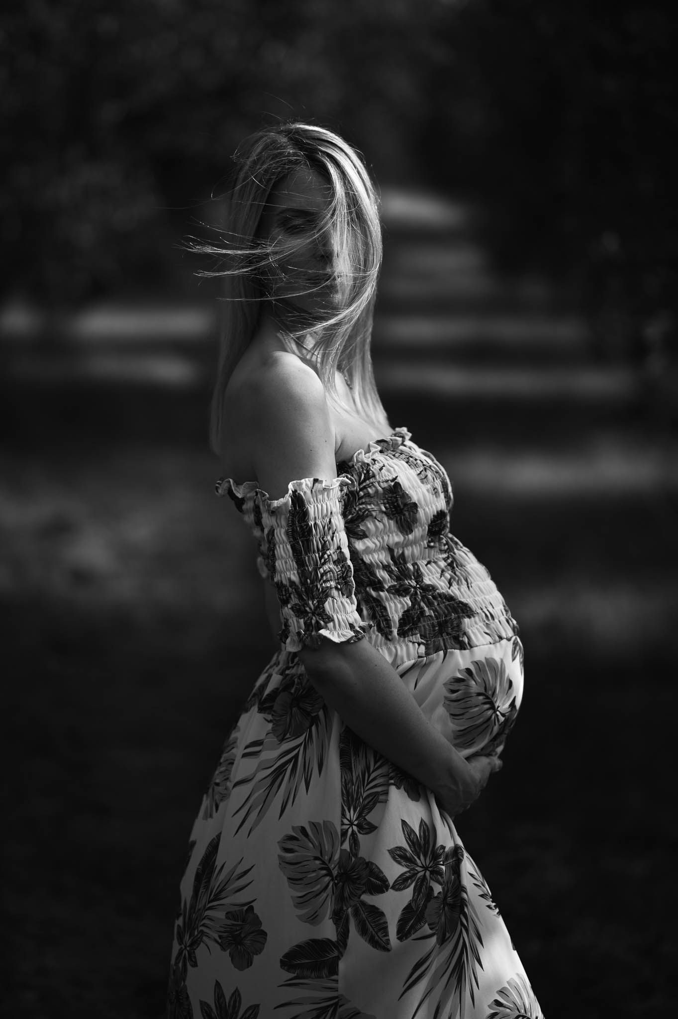 Black and white maternity photo of a blonde woman in a floral off-shoulder dress, cradling her baby bump outdoors.