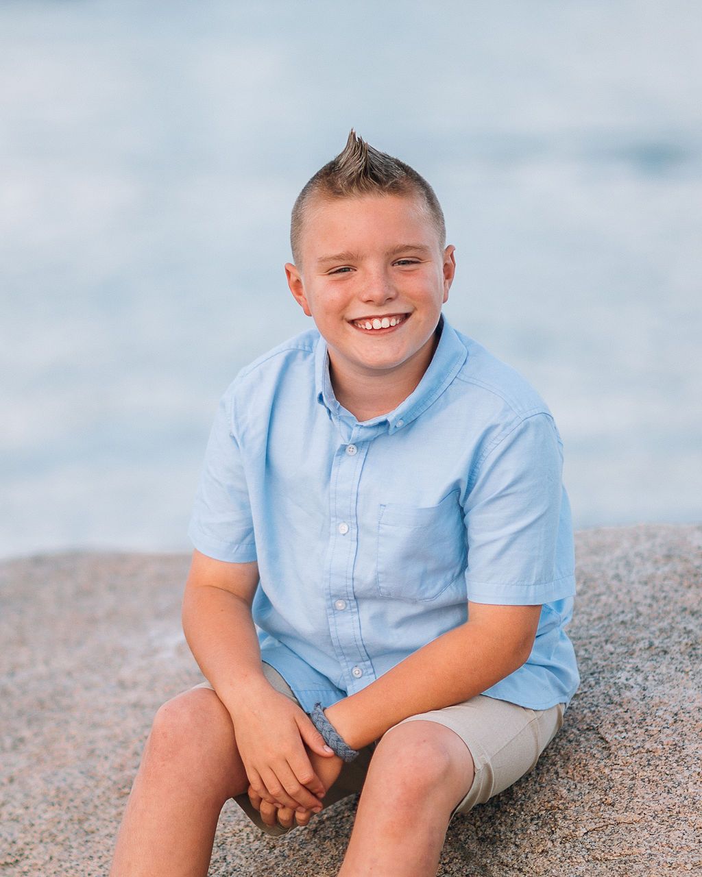 Smiling boy with a mohawk hairstyle wearing a light blue shirt, sitting on rocks near the ocean.