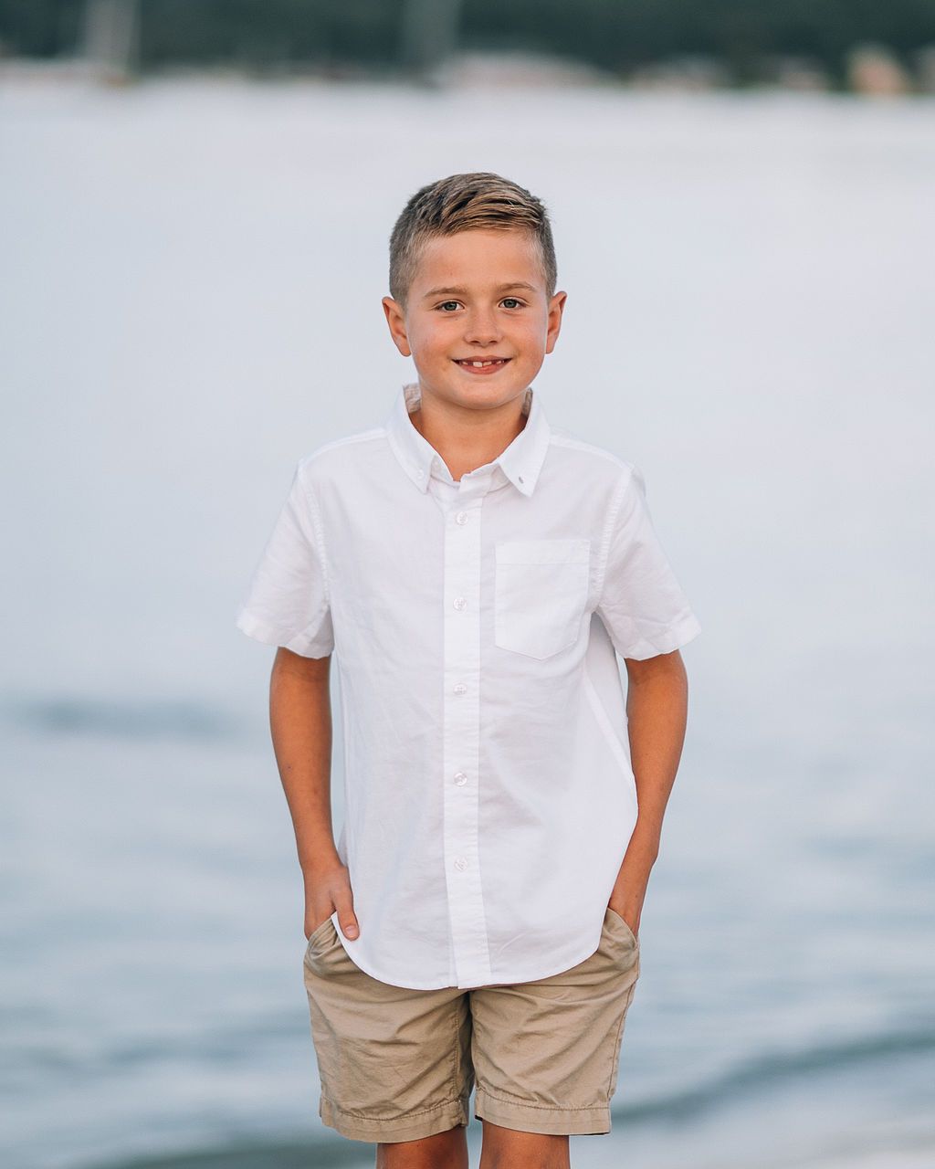 A smiling boy in a white button-up shirt and khaki shorts stands on a beach with water in the background.