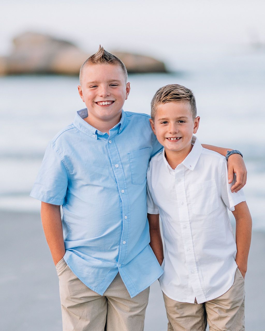 Two smiling boys in button-up shirts pose together on a sandy beach with ocean waves in the background.