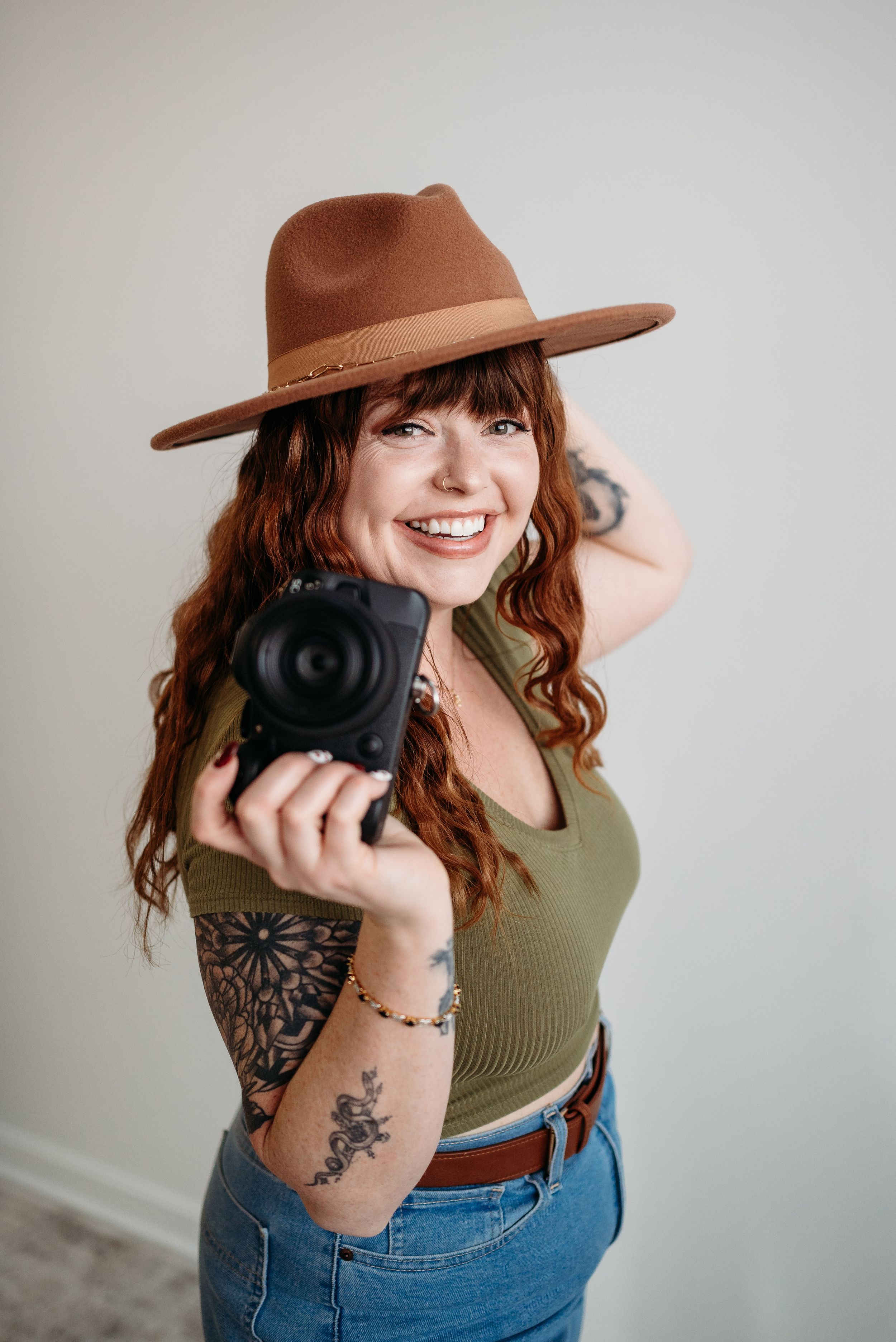 Smiling woman with red curly hair, brown wide-brim hat, tattoos, holding a camera, wearing green top and jeans.