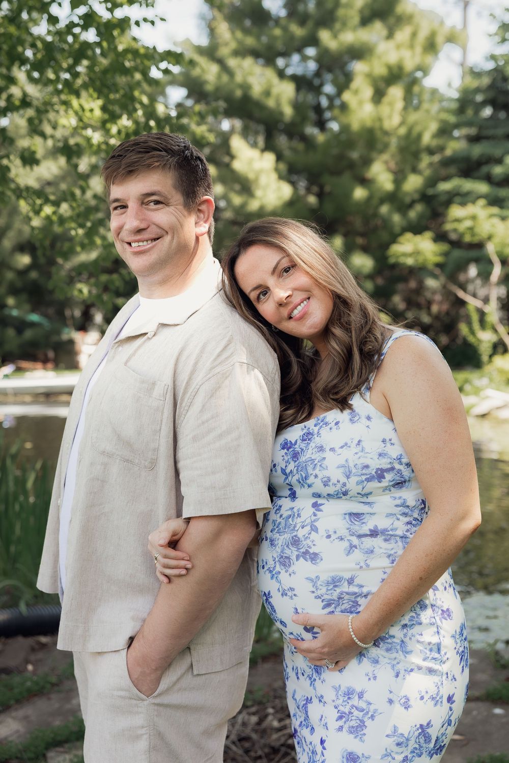 Expecting couple standing back-to-back outdoors, woman in blue floral maternity dress holding baby bump.
