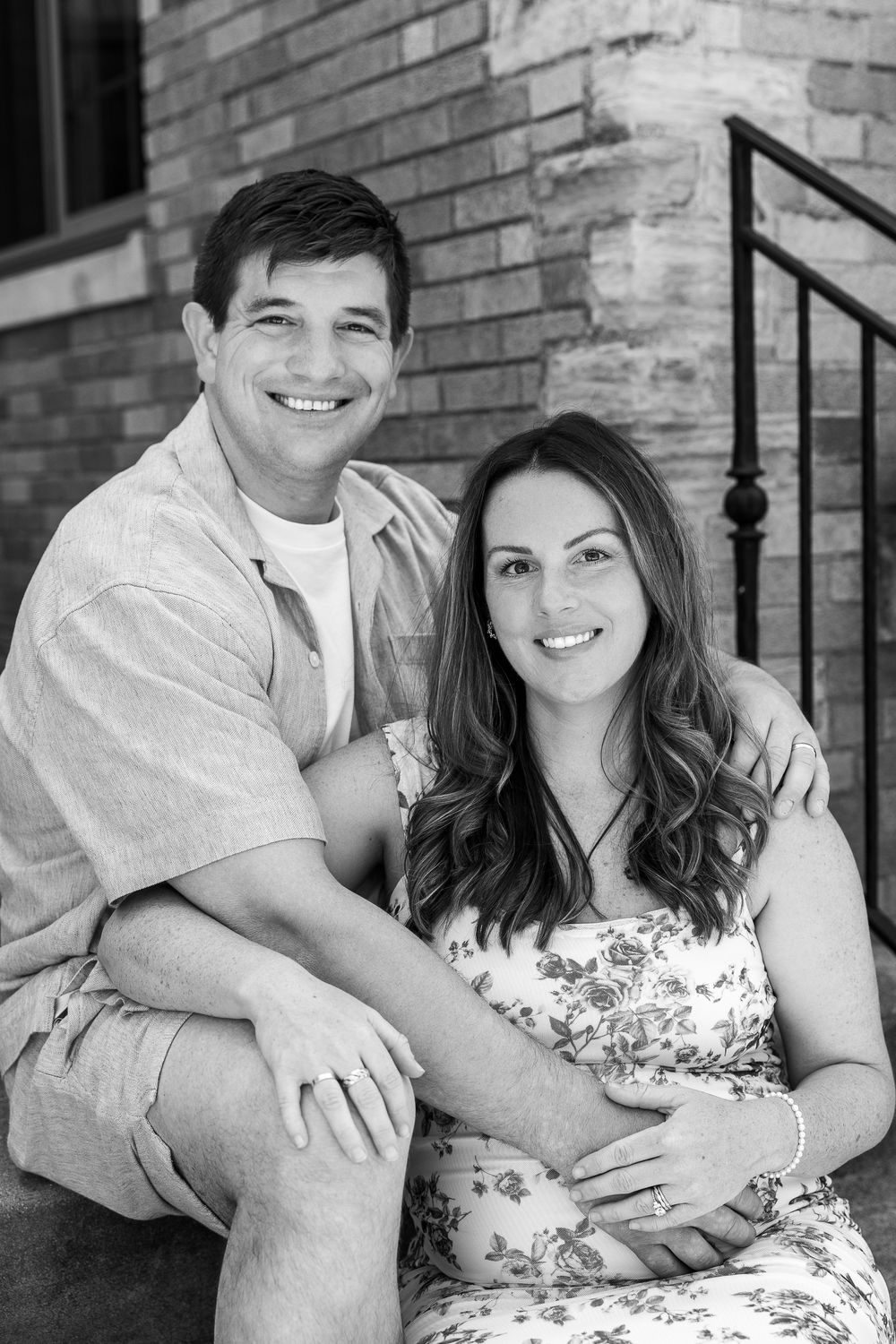 Black and white photo of a smiling couple sitting on steps in front of a brick building, woman in floral dress.