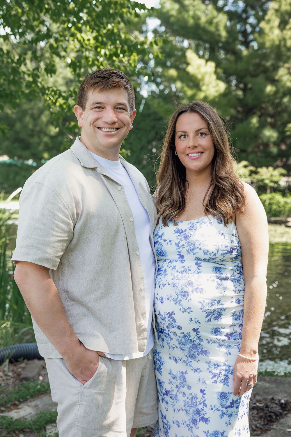 Smiling couple posing outdoors near a pond, woman in floral blue dress, man in beige linen shirt, lush green trees behind.