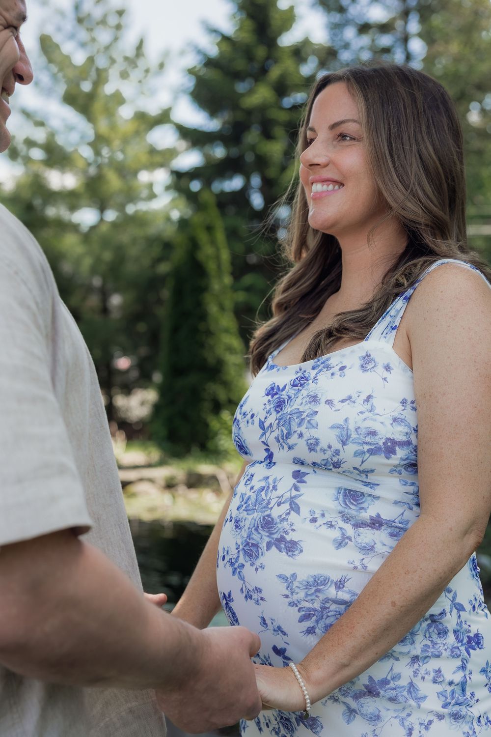 Smiling pregnant woman in blue floral dress holds hands with partner outdoors among green trees.
