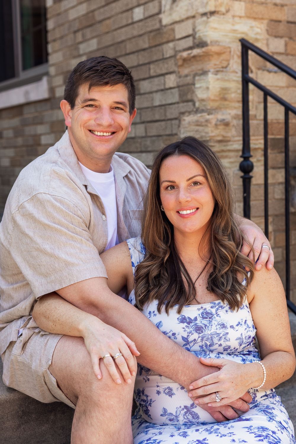 Smiling couple sitting on steps, pregnant woman in blue floral dress with partner in beige shirt.