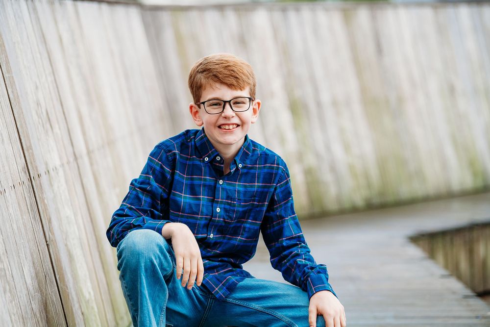 A red-haired boy wearing glasses and a blue plaid shirt sits smiling on an outdoor boardwalk.