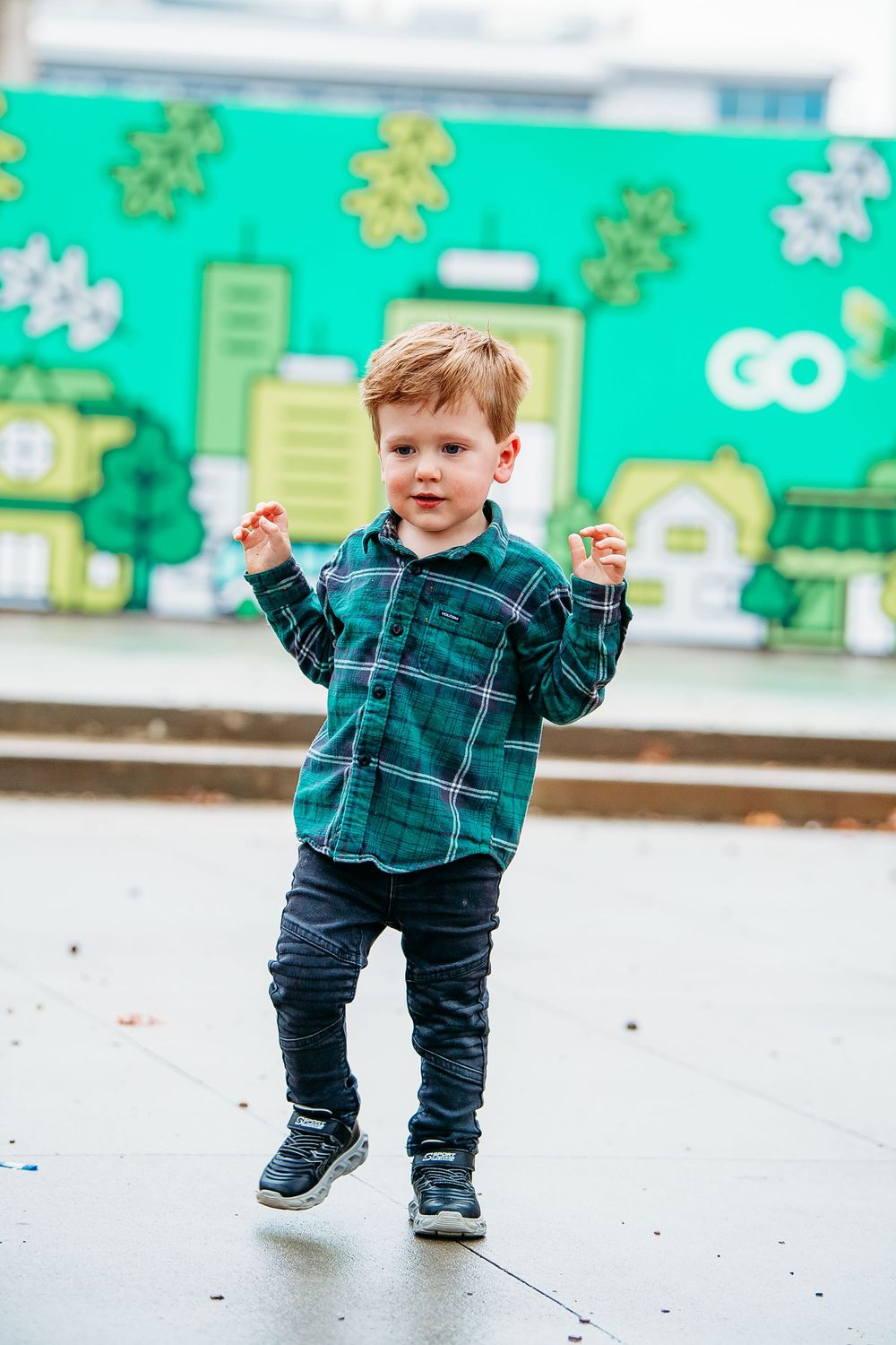 Toddler boy in green plaid shirt dancing outdoors in front of colorful green mural.
