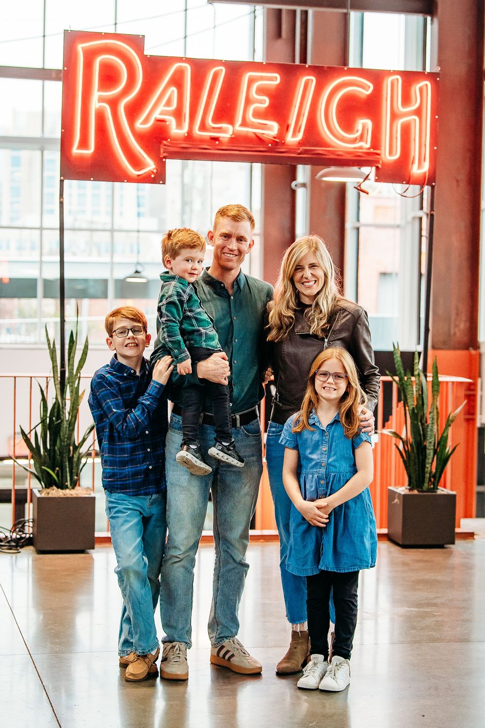 A family of five poses and smiles together under a glowing red Raleigh neon sign indoors.
