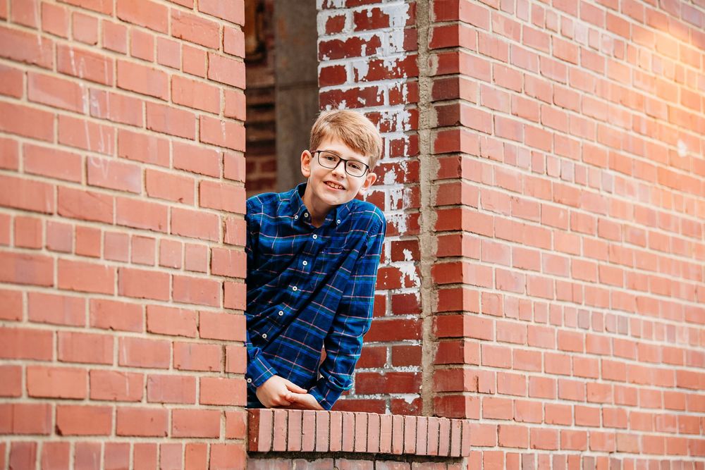 Young boy wearing glasses peeking through a rustic brick wall window opening.