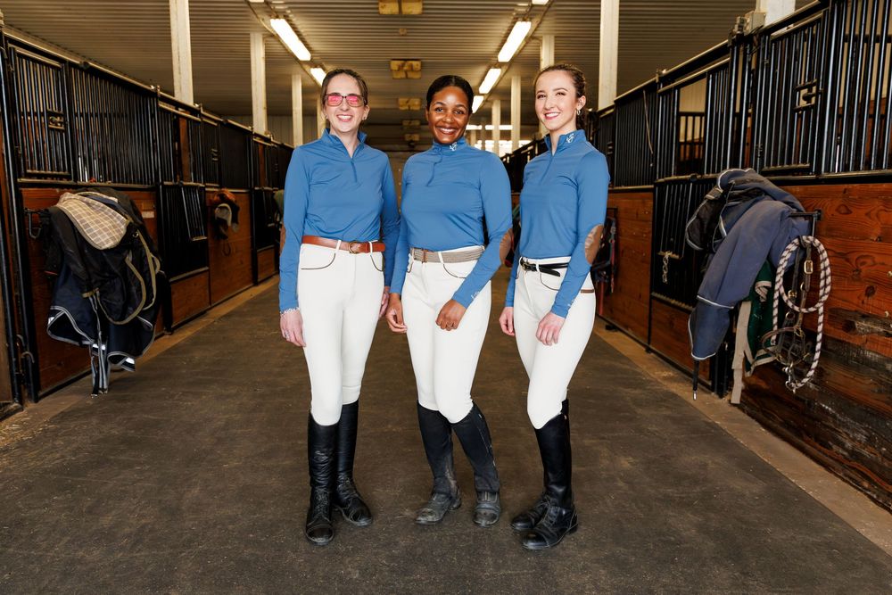 Three female equestrians in matching blue shirts and white riding breeches smiling in a horse stable.