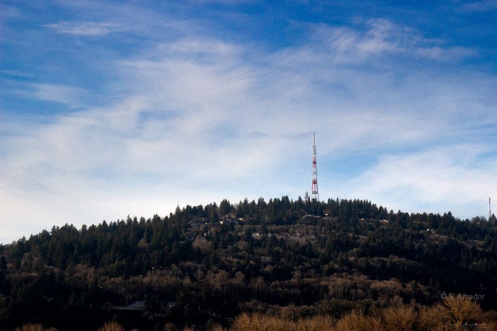 Forested hilltop with tall communication towers against a blue sky with wispy clouds.