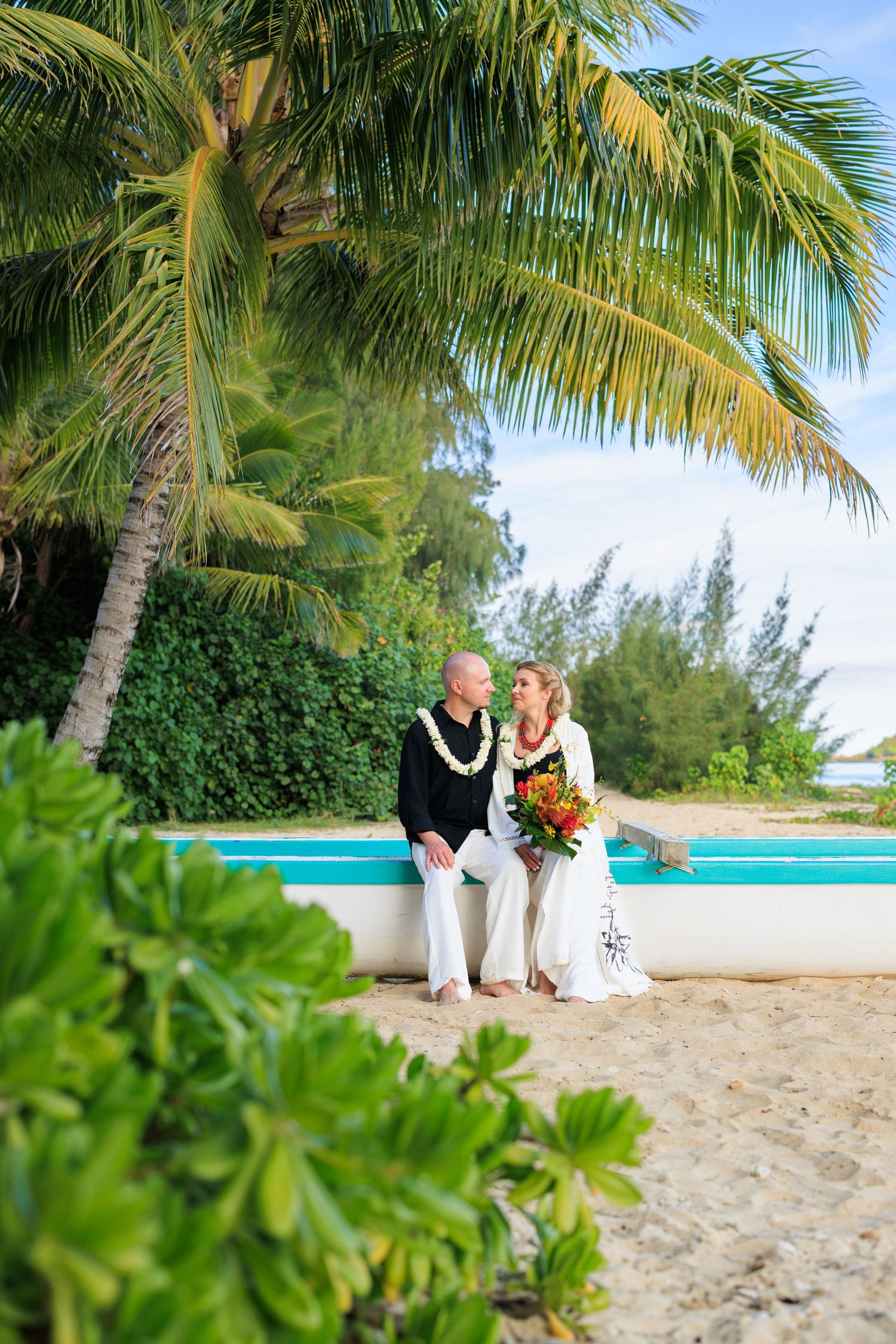 Couple in wedding attire sitting on a boat on a tropical beach with palm trees, bride holding a colorful bouquet.