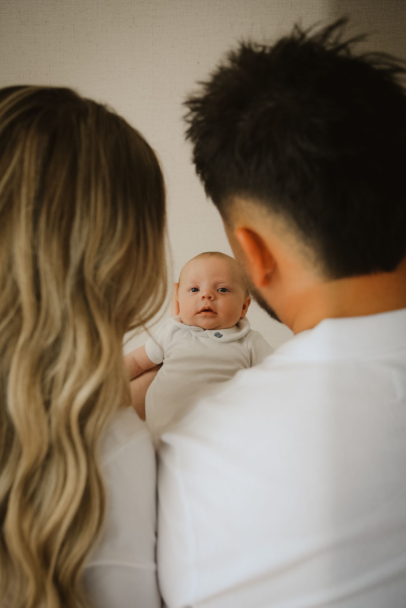 Parents holding their newborn baby in a white onesie, viewed from behind, with the infant gazing forward.