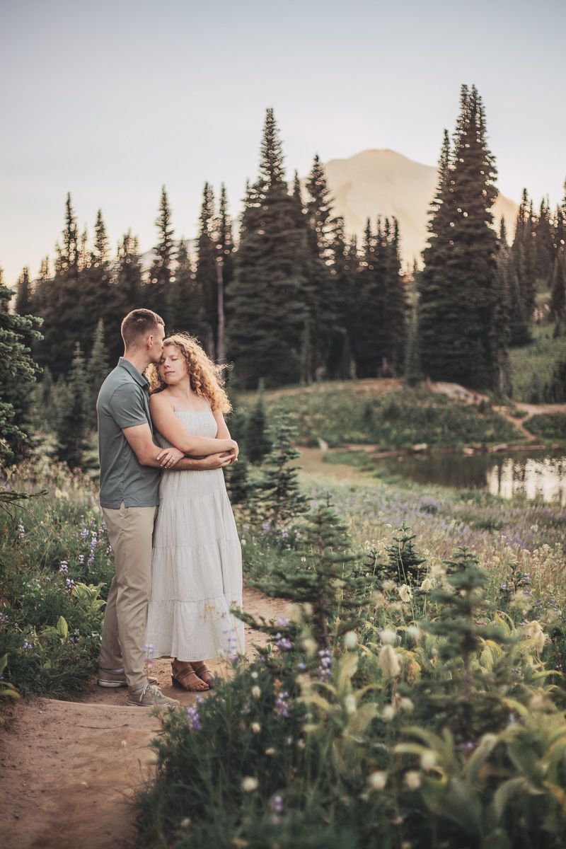 Couple embracing on a wildflower trail with a serene mountain lake and Mount Rainier behind them.
