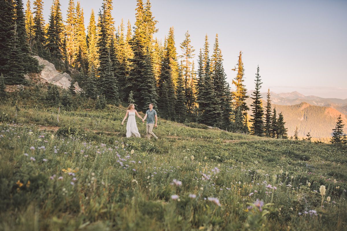 Couple holding hands walking through alpine wildflower meadow at sunset with tall evergreen trees.